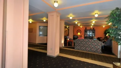 A living room or common area with orange walls, dark patterned carpet, and a couch where several people are seated