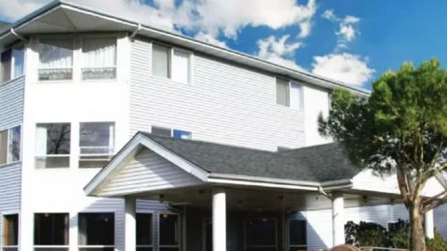 A three-story white building with a covered entrance and columns, with a large tree in front under a blue sky